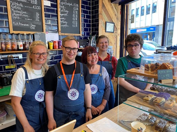 5 staff and supported volunteers at Artizan International - a charitable café that helps adults and young people with additional needs learn about employment - pose for a photo from behind the café counter.