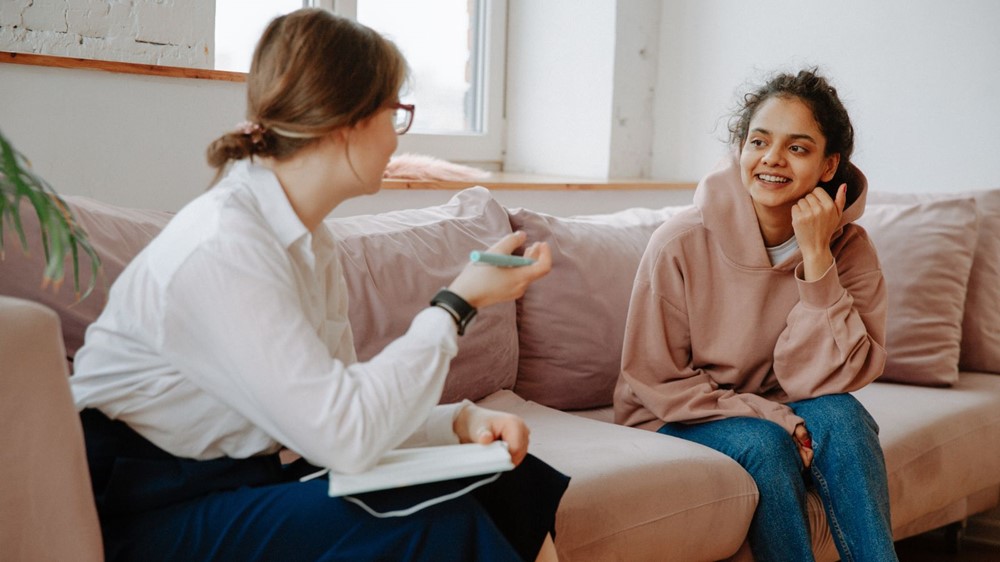 A female professional person sits on a sofa with a pen and paper, talking to a young adult female who sits on the sofa next to her.