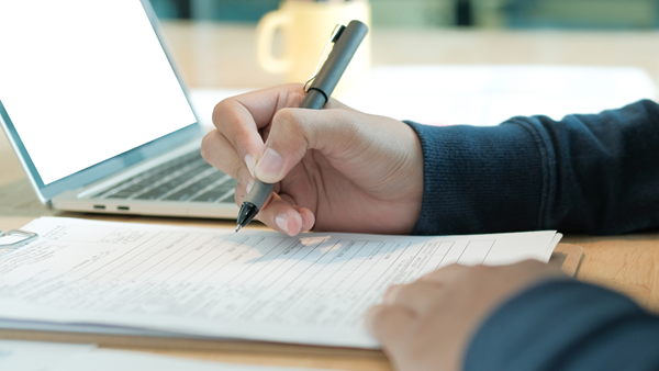 Close up photo of hands writing with a pen and paper next to an open laptop.